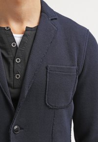 Blazer blu navy con texture, colletto a risvolto, taschino singolo e due bottoni, indossato sopra una camicia nera con bottoni.