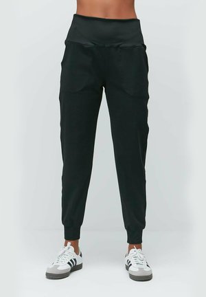 Schwarze Jogginghose mit hohem elastischen Bund, Seitentaschen, schmal zulaufenden Beinen und gerippten Bündchen. Kombiniert mit weißen Sneakers.