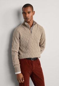 Beige Kabelstrickpullover mit einem Reißverschlusskragen, getragen über einem karierten Hemd mit Knöpfen. Kombiniert mit dunkelroten Cordhosen und einem schwarzen Gürtel.