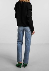 Schwarzes, tailliertes Oberteil mit strukturierten Puffärmeln, kombiniert mit blauen, geraden Jeans und grünen, hohen Mules. Einfacher, minimalistischer Designfokus.
