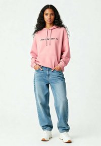 Felpa oversized rosa con tasca frontale e testo nero, abbinata a jeans baggy azzurri e sneakers bianche. Tessuto morbido.