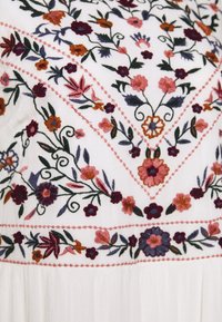 Un tessuto bianco ricamato presenta motivi floreali nei colori rosa, viola e arancione, con cuciture intricate e un aspetto testurizzato.