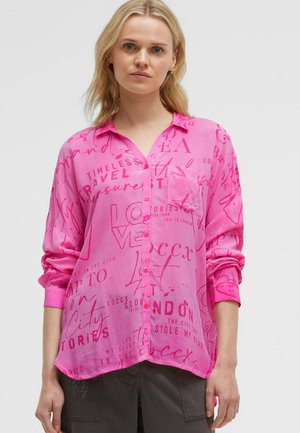 Blonde woman wearing a loose bright pink button-up shirt with red scripted text, paired with dark pants, against a plain gray background.