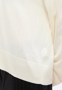 Gros plan sur l'ourlet d'un pull en maille crème avec bordure côtelée porté sur un pantalon noir à fines rayures.