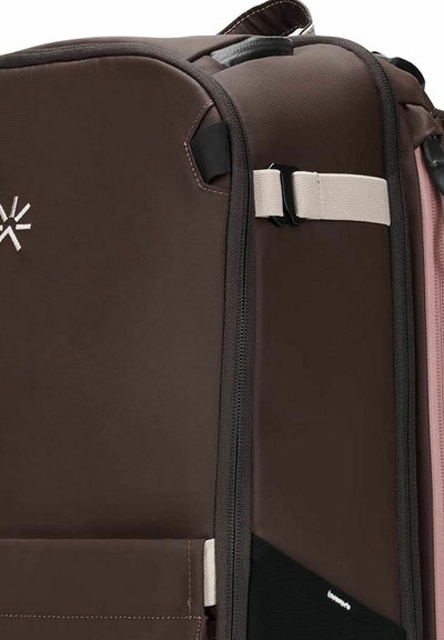 Brauner und schwarzer weich geformter Koffer mit Frontreißverschluss, beigem Riemen mit schwarzer Schnalle und einem kleinen weißen Logo auf der Stoffoberfläche.