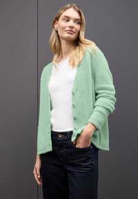 Mintgrön, fluffig cardigan med knappar framtill. Bärs över en vit skjorta, ihop med mörka jeans, som har en avslappnad passform och uppvikta ärmar.