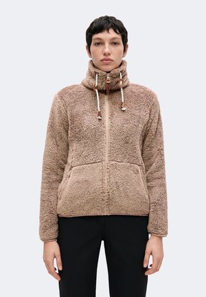 Personne portant une veste polaire beige zippée avec un col montant et des cordons, debout devant un fond clair uni.