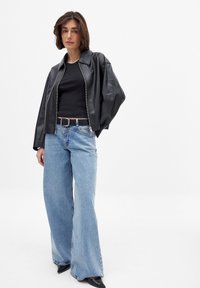 Juodas odinis užtrauktukas striukė su apykakle, derinama su plačiomis šviesiai mėlynomis džinsinėmis kelnėmis ir juodais smailiais bateliais.