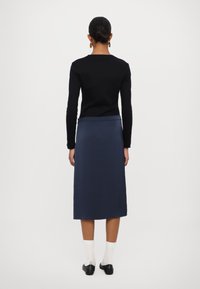 Melns garas piedurknes tops, pieguļošs viduklī; tumši zils midi svārki, gluda tekstūra; saskaņots ar baltām zeķēm un melniem balerīniem, redzams no aizmugures.