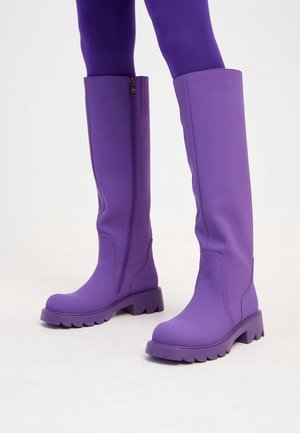 Bottes hautes violettes avec semelles épaisses portées par-dessus des collants violets assortis, présentées sur un fond clair uni.