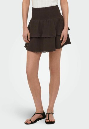 Mujer con una mini falda negra de capas, top blanco sin mangas y sandalias planas negras, de pie frente a un fondo liso.