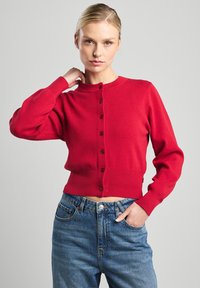 Gilet rouge court à manches longues, texture côtelée, encolure ronde et fermeture à six boutons. Associé avec un jean taille haute bleu.