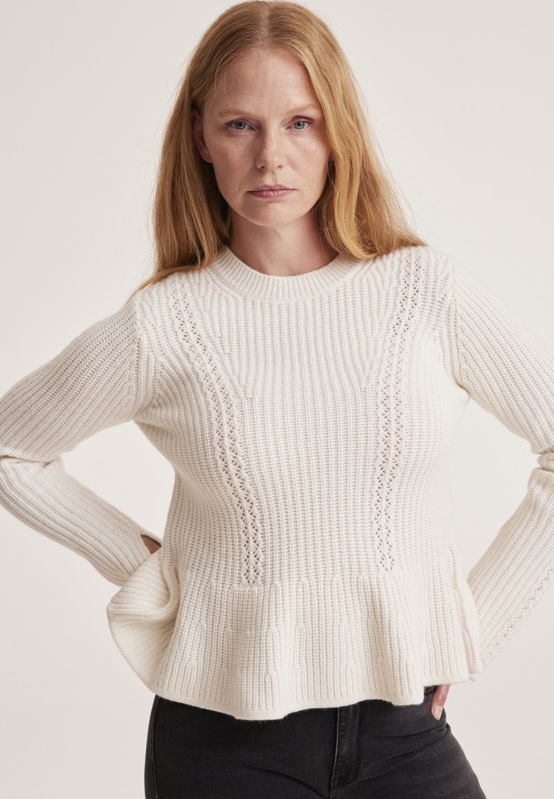 White knitted sweater with a ribbed texture, peplum hem, and lace details along the sides. Features a crew neckline and long sleeves.