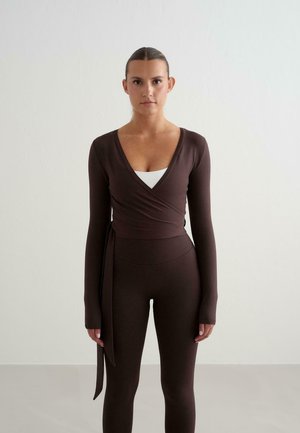 Woman with hair tied back wearing a dark brown wrap long-sleeve top over a white tank and matching dark brown leggings, standing against a plain background.
