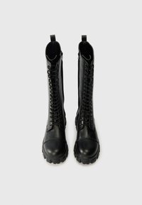 Μαύρες δερμάτινες μπότες μέχρι το γόνατο με στρογγυλή μύτη, κορδόνια μπροστά και αντίθετους χοντρές σόλες. Παρουσιάζουν λεία υφή και θηλιά για εύκολη τοποθέτηση στην κορυφή.