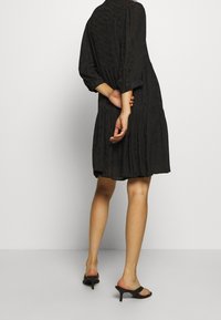 Vestido negro con mangas tres cuartos, tela texturada y diseño en capas. Se lleva con sandalias negras de tacón, mostrando las piernas y los zapatos.