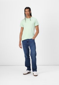 Jasnozielona bawełniana koszulka z okrągłym dekoltem i krótkimi rękawami, zestawiona z niebieskimi dżinsami i białymi sneakersami z brązowymi podeszwami.