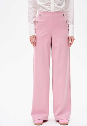 Różowe spodnie o szerokich nogawkach z gładką teksturą, wysoki stan i metalowe guziki po bokach. Zestawione z białą koszulą na guziki.