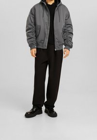 Grå vadderad bomberjacka med huva, med elastiska manschetter och sidofickor, bärs över en svart stickad tröja och lösa svarta byxor.