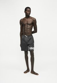 Homme grand, musclé et à la peau foncée, debout pieds nus, portant un short noir à motifs avec le logo blanc "DSQ" sur la jambe gauche, les mains derrière le dos.