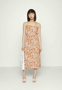 Mulher em pé, usando um vestido midi castanho com padrões abstratos brancos, segurando uma mala branca e usando sandálias brancas em slide.