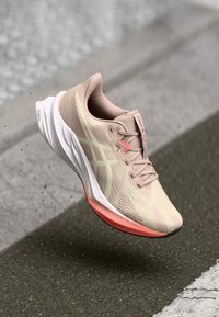 Hardloopschoen met een lichtbeige textiele bovenkant, witte middenzool en een roze-oranje buitenzool. Voorzien van textuurpatronen en een vetersysteem.