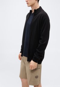 Μαύρη ζακέτα με φερμουάρ και υφή, σε συνδυασμό με μπλε κοντομάνικο πουκάμισο και μπεζ σορτς με πλαϊνή τσέπη και λογότυπο.