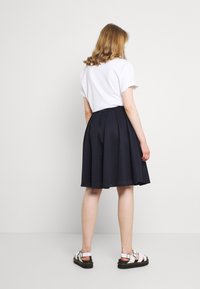 Marinblå plisserad kjol med en strukturerad design, kombinerad med en vit kortärmad skjorta och vita sandaler på en modell som står framför en enfärgad bakgrund.