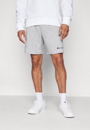 Mężczyzna ubrany w szare szorty marki Champion, białe skarpetki, białe trampki oraz białą bluzę, stojący na jednolitym jasnym tle.