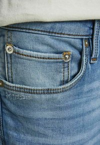 Gros plan sur une poche de jean bleu en denim avec des rivets métalliques et des coutures visibles, portée avec un tissu blanc rentré au-dessus.