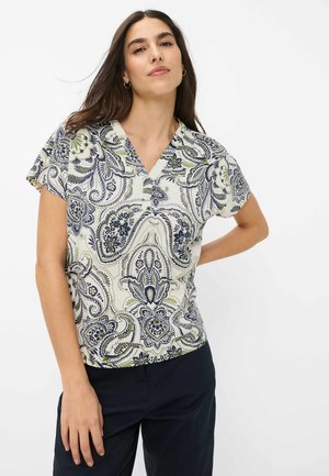 Kvinde iført bluse med korte ærmer og V-udskæring med marineblåt og grønt paisley-mønster, kombineret med mørke marineblå bukser, står mod en hvid baggrund.
