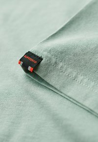 Tessuto verde menta con una texture morbida. Etichetta nera con "SUPERDRY" in accenti arancioni e rossi cucita sul bordo. Dettagli delle cuciture ondulate visibili.