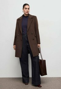 Cappotto doppiopetto in misto lana marrone con colletto a revers, abbinato a pantaloni larghi scuri a gamba larga. Completo di borsa shopper marrone e scarpe con tacco.