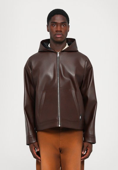 Jeune homme portant une veste à capuche zippée marron foncé avec des poches avant et un pantalon orange brûlé, se tenant devant un fond uni.