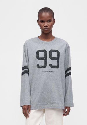 Personne portant une chemise grise à manches longues avec le numéro noir 99 et deux bandes noires sur chaque manche, associée à un pantalon clair.