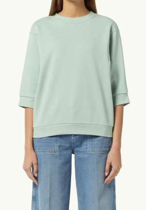 Femme portant un sweat-shirt oversize à manches courtes vert clair et un jean taille haute bleu clair, debout devant un fond uni.