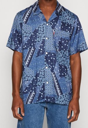 Chemise à manches courtes boutonnée avec un motif patchwork bleu comprenant des dessins floraux et géométriques, portée avec un jean en denim bleu.