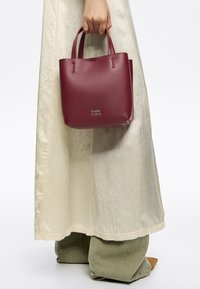 Torebka Bimba y Lola z bordowej skóry, o wyraźnej konstrukcji, z dwoma krótkimi uchwytami, logo z przodu, umieszczona na jasnej, fakturowanej sukience.