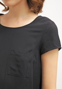 Czarna bluzka z krótkim rękawem o okrągłym dekolcie i kieszonką z przodu. Gładka tekstura i luźny krój.