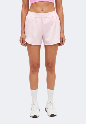Vrouw die lichtroze sportshorts, witte sneakersokken en witte sneakers draagt, staande tegen een eenvoudige lichte achtergrond.