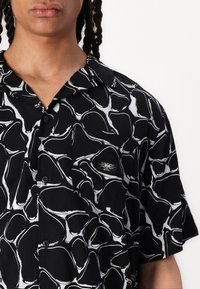 Camicia nera a maniche corte con un motivo astratto bianco, chiusura frontale con bottoni, ampio colletto e una piccola etichetta con logo sulla tasca sinistra del petto.