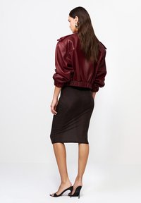 Kobieta z długimi włosami, ubrana w burgundową skórzaną kurtkę, czarną ołówkową spódnicę i czarne sandały na wysokim obcasie, stojąca plecami.