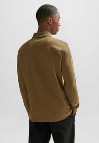 Bruine poloshirt met lange mouwen en een platte kraag, zachte textuur en zijsplitten, gezien vanaf de achterkant. Draagt donkere broek.