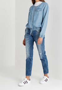 Chaqueta de mezclilla azul claro con dos bolsillos en el pecho, cierre de cremallera y cintura ajustable con cordón, combinada con jeans azul desgastados y zapatillas blancas.