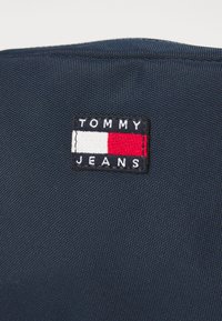 Jūras zila auduma ar teksturētu virsmu, kurā ir taisnstūra izšuvuma plāksteris. Uz plākstera ir balts uzraksts "TOMMY JEANS", ko ieskauj sarkans un balts svītrojums.