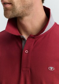 Camisa polo roja con cuello gris y acentos grises, fabricada con un tejido texturizado y con una tapeta de dos botones. Pequeño logo en el pecho.