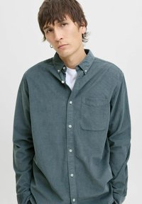 Grønn kordfløyelskjorte med knappet krage, enkelt brystlomme og subtile vertikale striper, båret over en hvit T-skjorte.