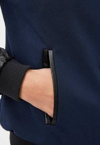 Chaqueta azul marino con un tejido texturizado, acento de cuero negro en el bolsillo y puños acanalados. Presenta una mano descansando en el bolsillo lateral.
