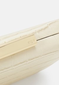 Käsilaukku vaalean kermanväristä teksturoitua keinonahkaa, jossa on kullanvärinen metallilukko kaiverretulla logolla ja hienovaraisesti krokotiilikuosisella pinnalla.