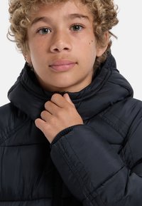 Ung person med lockigt blont hår som bär en svart puffjacka, håller dragkedjan nära halsen och ser rakt fram.